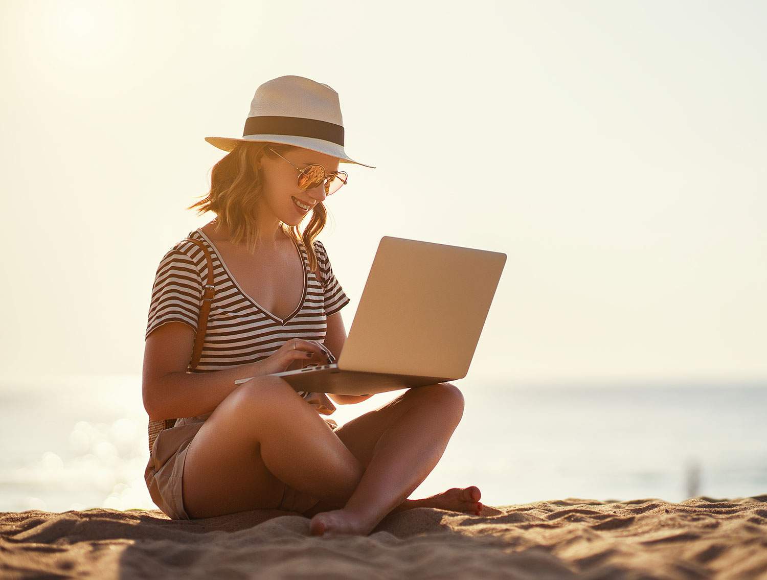 Woman sitting in the sand by the beach, using a laptop - how much commission do travel agents make?