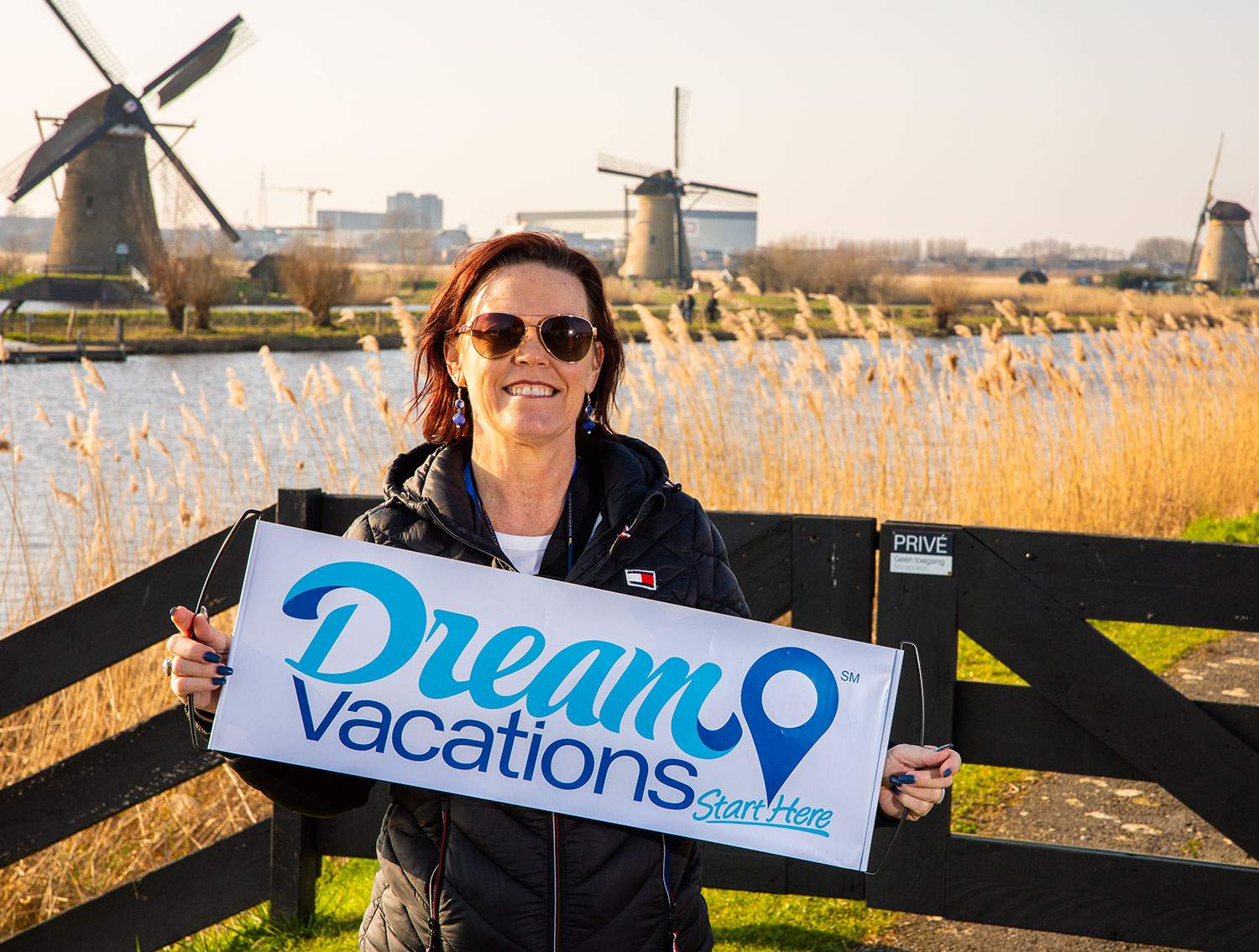 A woman standing near a couple windmills in a field, holding a Dream Vacations sign