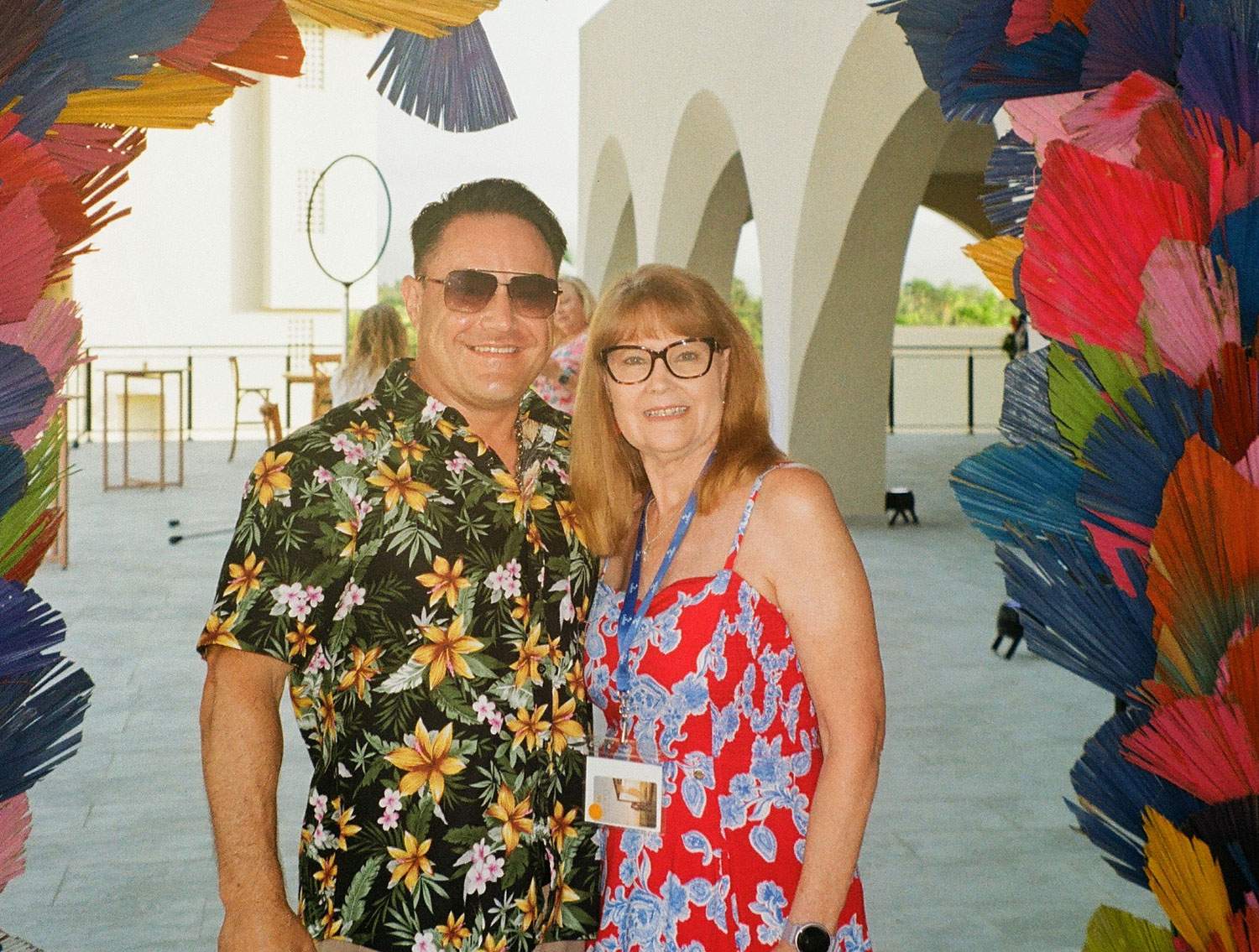 A man and woman stand together in front of a vibrant, colorful archway, smiling at the camera.